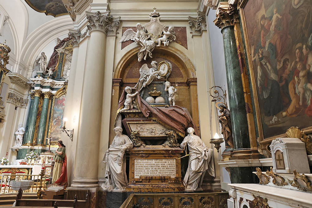santa barbara-029 A la derecha del altar se halla el sepulcro de pórfido y mármol del Rey Fernando VI ejecutado por Francisco Gutiérrez (1724/1782) siguiendo diseños de Francisco Sabatini (1721/1797) (fig 7) que se considera una de las obras cumbre del Neoclasicismo español. En su parte superior resalta una escena tallada de Su Majestad protegiendo las artes y a sus lados, simbolizando las virtudes de su reinado, alegorías de la abundancia y la justicia. Corona la obra una representación de Saturno, como dios del tiempo, acentuando el peso de lo inexorable. En la parte inferior se lee una inscripción latina que se traduce así: “Aquí yace el fundador de este monasterio Fernando VI, Rey de las Españas, óptimo príncipe que murió sin hijos, pero con numerosa prole de virtudes. Padre de la Patria, el 10 de agosto de 1759. Carlos III dedicó este monumento de tristeza y piedad a su queridísimo hermano, cuya vida hubiera preferido al Reino